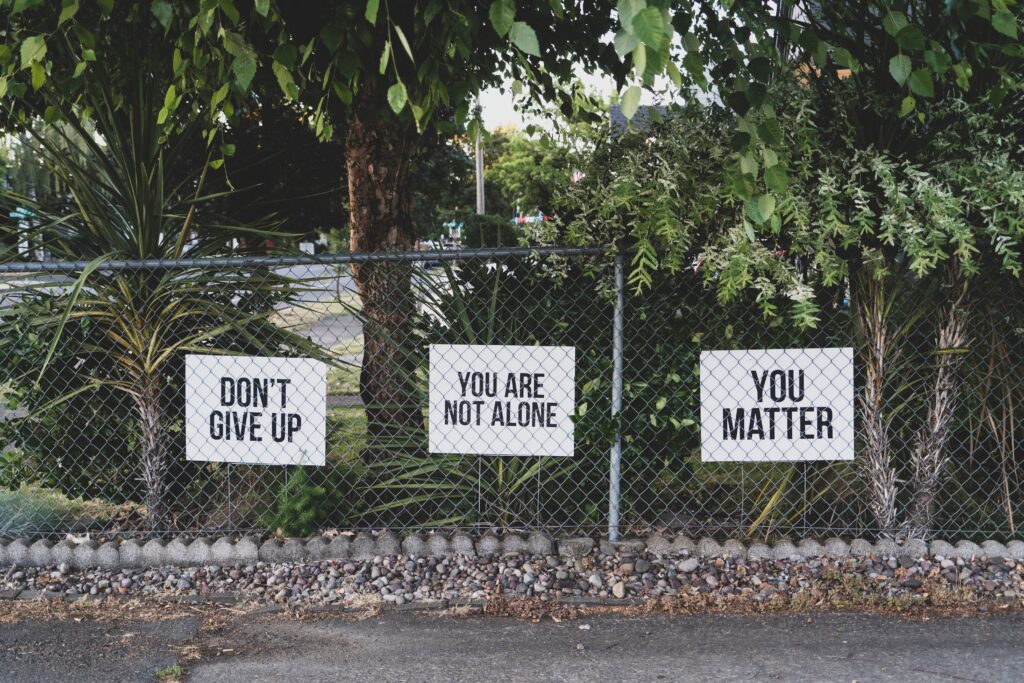 A stretch of chainlink fence in a city, with three sign encouraging mental health taped to the fence. They say "Don't Give Up," "You Are Not Alone," and "You Matter."