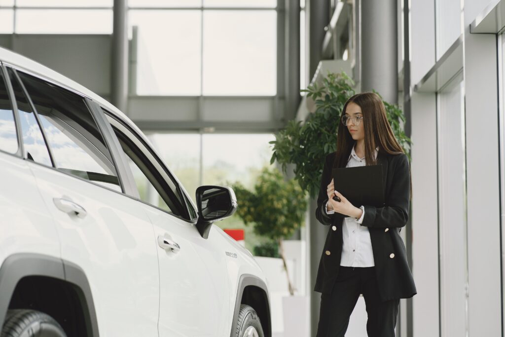 A young woman holding a clipboard, looking at a car in a dealer showroom with a quizzical look.